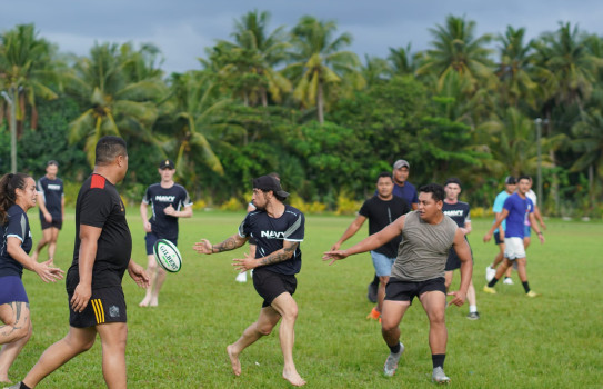 Navy personnel compete in a friendly game of rugby with the Samoan Police Force in front of palm trees. Team Navy are wearing PT uniform and some are playing barefoot.