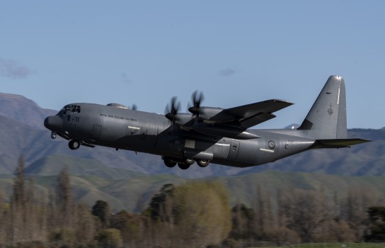 A C-130J Hercules in the air on a sunny day with green hills in the background - blue sky. 