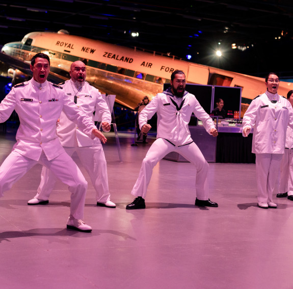 5 sailors wearing white formal uniform perform a haka in front of an ex-RNZAF aircraft.