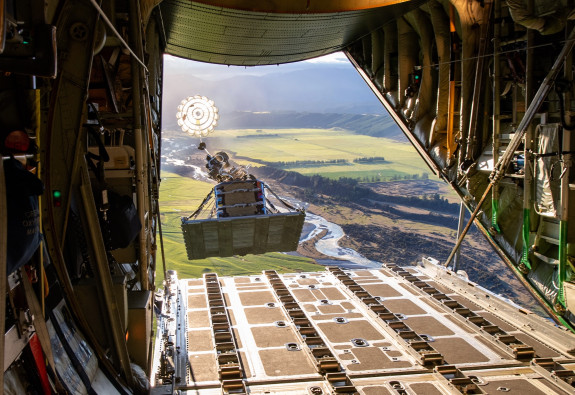 A large wooden crate called a "load" with a open parachute attached to it falling outside of the back of an Hercules aircraft.