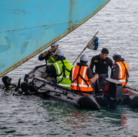 Navy and Customs personnel drifts next to a large container ship on a Rigid Hull Inflatable Boat. Two divers in the water talk with them about the search.