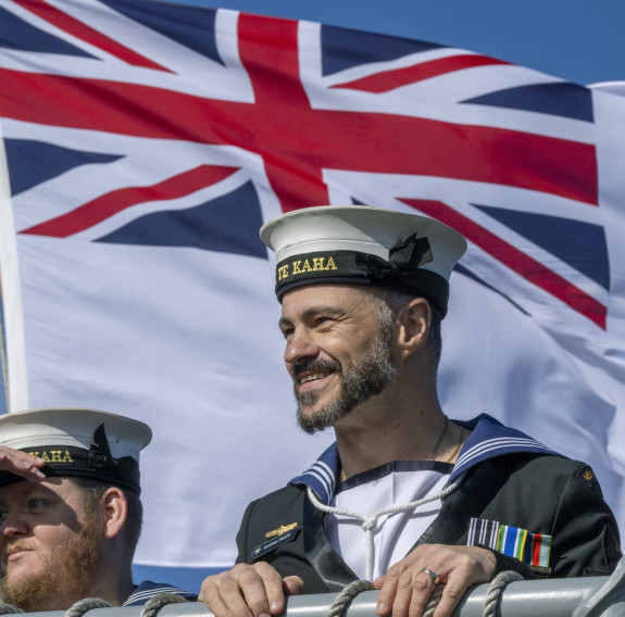 A sailor wearing the Navy uniform smiles as the ship arrives. The Navy's flag blows in the wind behind him.