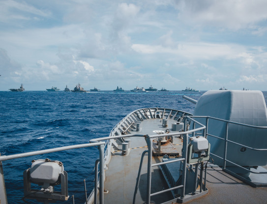 The bow of HMNZS Te Kaha on the sea and its 5-inch gun, with a formation of foreign military vessels in the background.