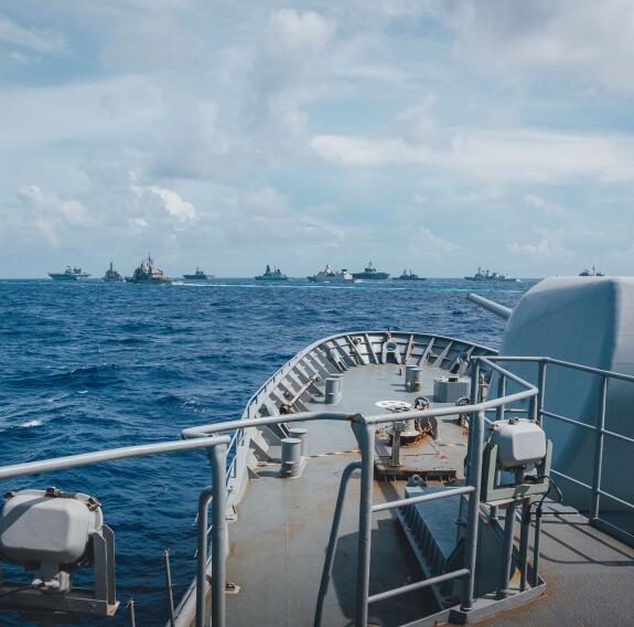 The bow of HMNZS Te Kaha on the sea and its 5-inch gun, with a formation of foreign military vessels in the background.