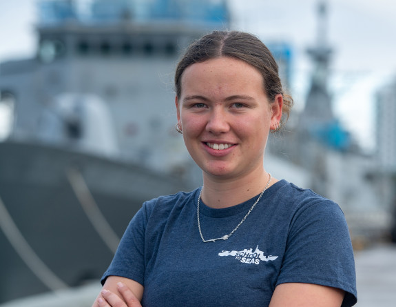 Thaeya Hoyle, a School to Seas participant stands in front of a RNZN ship, she is wearing a blue 'School to Seas' branded t-shirt and a necklace.