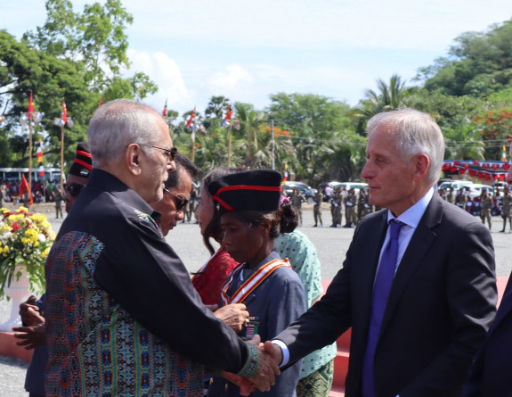 Two men shake hands in the bright daylight, people congregate in the area.