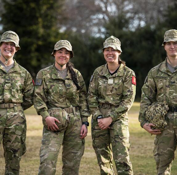 Four soldiers stand together, each wearing camouflage uniform and hats, in front of a field and trees in the background.