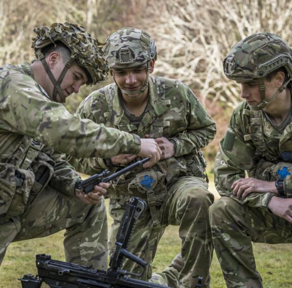 Three soldiers kneel while assessing part of a weapon. The soldiers are in full uniform with large belts on and helmets.