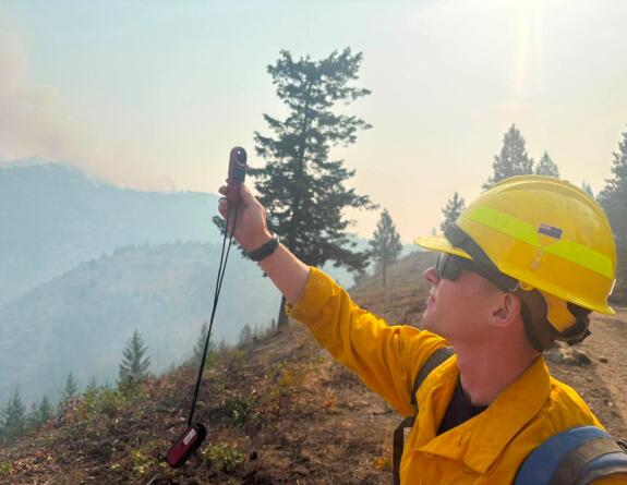A person stands with a uniform on and yellow hard hat with their right arm up as they reads a weather meter. In the background is a tree and it is smoky.