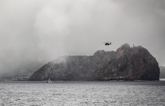 A Royal New Zealand Air Force NH90 helicopter in the air near the Whaakari/White Island. In the image you can see smoke erupting from the volcano.