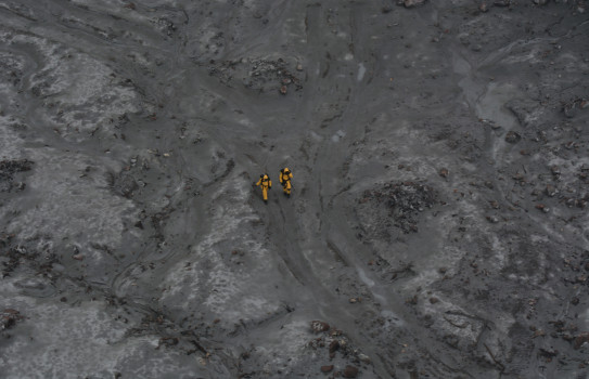 Two New Zealand Defence personnel wearing yellow suits walk on grey volcanic terrain on White Island