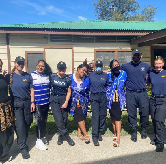 Navy stewarts with students and staff of the Ahopanilolo Technical Institute pose wearing a variety of blue uniforms, they stand in front of one of the facilities buildings and blue sky.