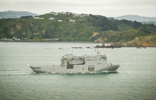 A grey ship sails past rocks and a hill with scattered houses on it.