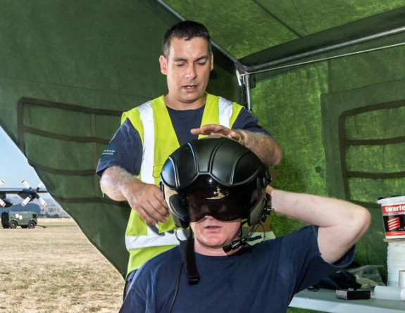 A Royal New Zealand Air Force Safety and surface technician checks the helmet on an airmen in a tent with the C-130H(NZ) Hercules aircraft on the flightline in the back on a nice day