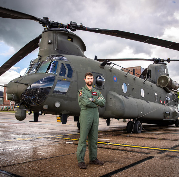 A man stands in green overalls with his arms crossed in front of a Royal Air Force Chinook helicopter on an overcast day.