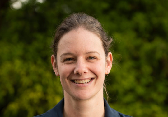 Flight Lieutenant Nina White in operational uniform, smiling and crossing her arms in a relaxed manner. A lush green landscape backdrops her.