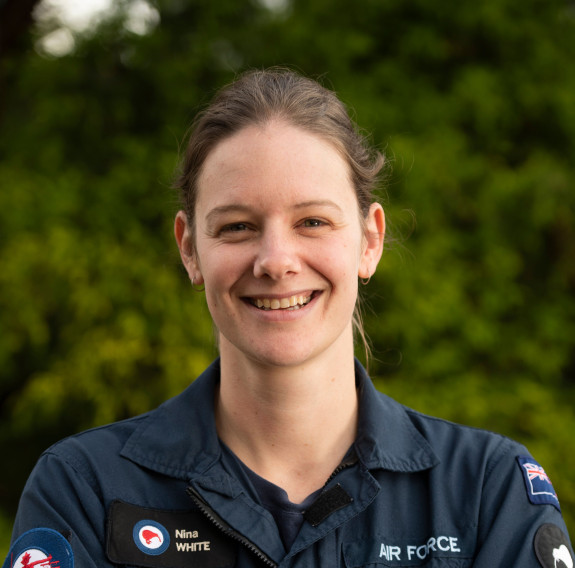 Flight Lieutenant Nina White in operational uniform, smiling and crossing her arms in a relaxed manner. A lush green landscape backdrops her.