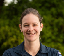 Flight Lieutenant Nina White in operational uniform, smiling and crossing her arms in a relaxed manner. A lush green landscape backdrops her.