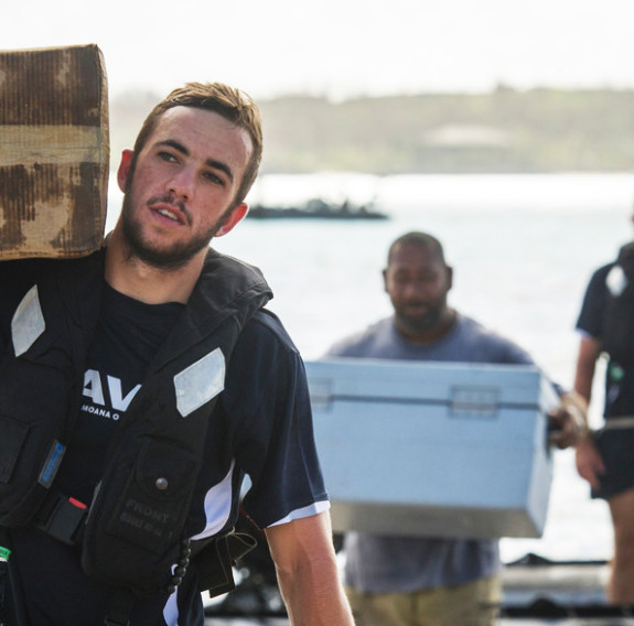 A Navy sailor in line with sea in background, holding a box of supplies on their shoulder for delivery to Fiji.