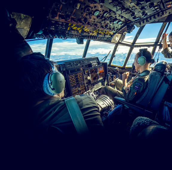 Inside the cockpit of a C-130 hercules, three aircrew members with headset on navigate a blue and cloudy sky