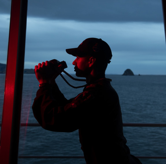 A Royal New Zealand Navy sailor on the bridge of a ship about to look through some binoculars. It's really dark, and you can see the red light from the bridge. The sailor is looking to the left of the picture and to their right, is the ocean and the bridg
