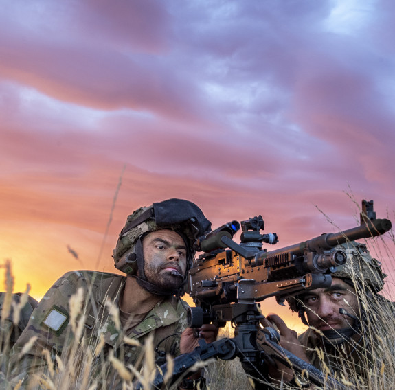At dusk two soldiers are in position, laying between dry grass, holding a large machine gun while looking into the distance for enemy movement