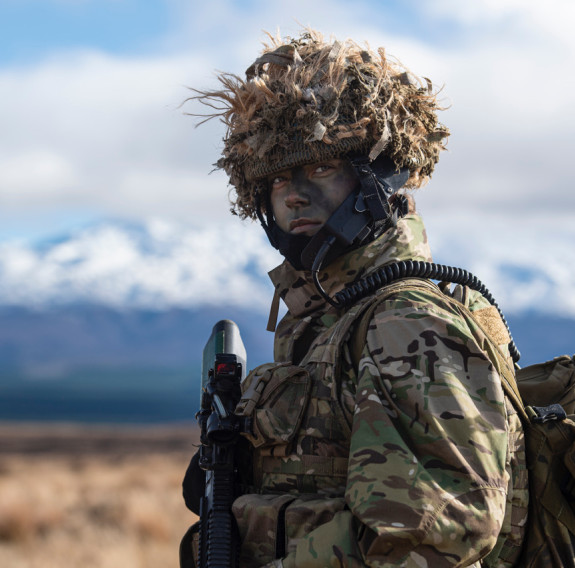 A New Zealand Army soldier stands with full uniform and headgear and a weapon. In the background, you can see snow-capped Mt Ruapehu