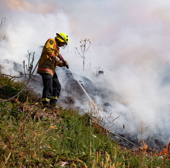An NZDF firefighter hoses down smouldering and burning grass on a hill in Nelson. 