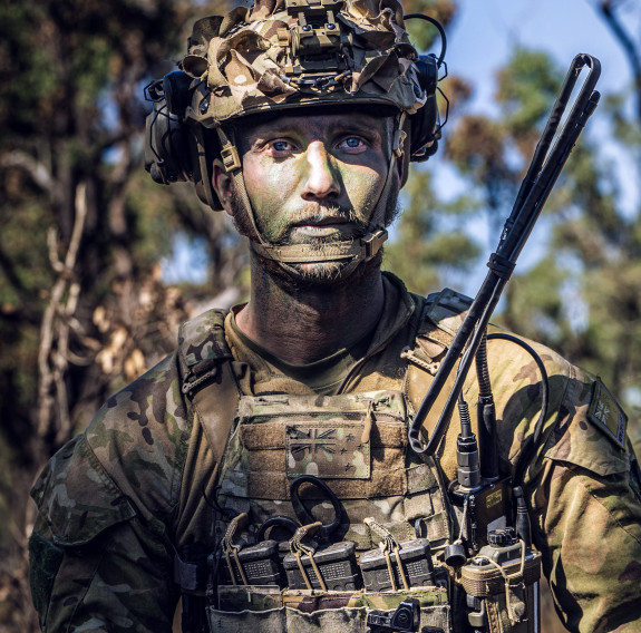 A soldier in uniform looks to their left. They are in the bush, trees in the background and blue sky. They have a helmet on and camo paint. 